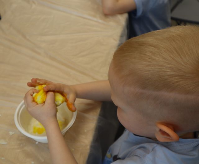 Na zdjęciu znajduje się chłopiec przy stole ugniatający w rękach żółtą ciastolinę. Zdjęcie zostało wykonane od góry.