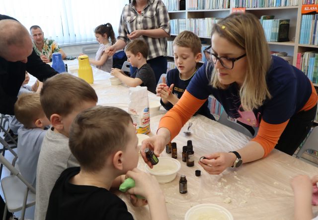 Na zdjęciu znajdują się dzieci przy złączonych stołach, oraz trójka dorosłych, jedna kobieta się nachyla i daje chłopcu do powąchania zapach. W tle znajdują się okna oraz półki z książkami.