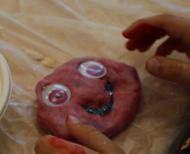 Na zdjęciu znajduje się różowa ciastolina z oczami i uśmiechem, leżąca na folii na stole, po obu jej stronach znajdują się dziecięce ręce.