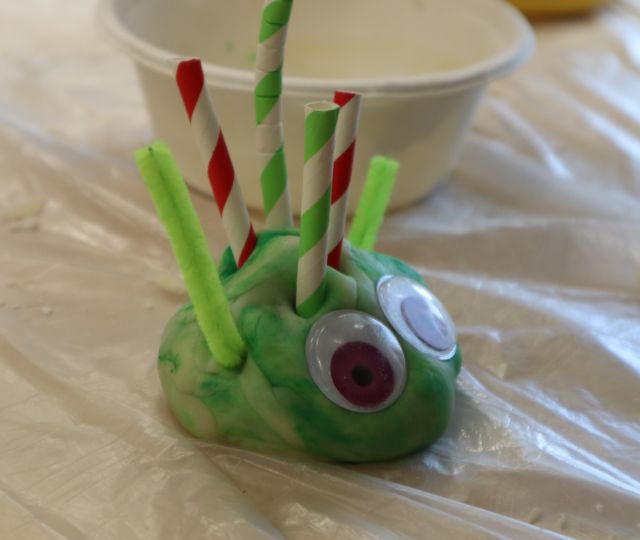 Na zdjęciu znajduje się zielona ciastolina z oczami i kawałkami słomek. Za pracą znajduje się miseczka.