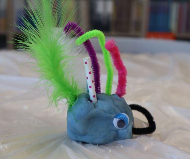 Na zdjęciu znajduje się leżąca na stole owiniętym folią niebieska ciastolina z oczami, kawałkami słomek, zielonym piórkiem i drucikami kreatywnymi. W tle znajdują się półki z książkami.