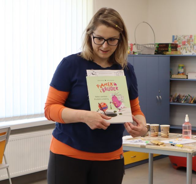 Na zdjęciu znajduje się kobieta, która trzyma w rękach książkę, za kobietą w tle znajduje się stolik, szafki i okna.