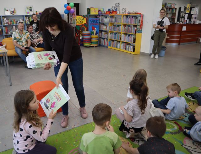 Na zdjęciu znajdują się siedzące na dywanie dzieci, a przed dywanem stoi kobieta z książkami i daje każdemu uczestnikowi po jednej. W tle znajdują się półki z książkami oraz rodzice.