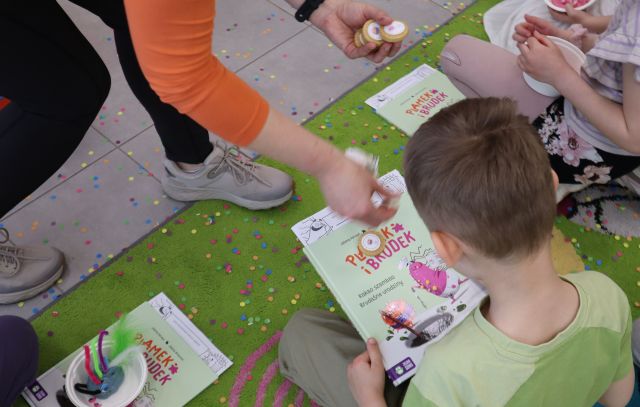Na zdjęciu znajduje się trójka dzieci siedząca na dywanie z książkami, a kobieta przed nimi daje im po mydełku. Zdjęcie zostało wykonane od góry.