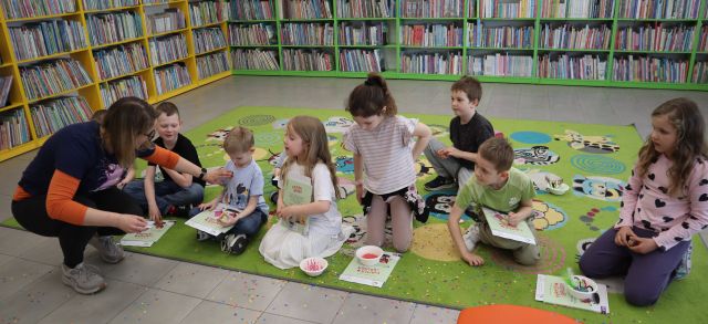 Na zdjęciu znajduje się ośmioro dzieci na dywanie, a przed nimi nachyla się kobieta i przykleja każdemu na koszulkę naklejkę. W tle znajdują się półki z książkami.