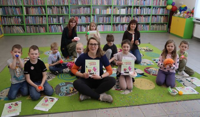 Na zdjęciu znajduje się dziewięcioro dzieci siedzących na dywanie do zdjęcia wraz z książkami, wraz z nimi siedzą jeszcze trzy kobiety. W tle znajdują się półki z książkami.