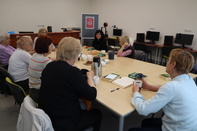 Na zdjęciu znajdują się kobiety siedzące przy złączonych stołach, mają przed sobą książki, a na stole znajduje się proporczyk oraz termosy i szklanki. Warsztaty odbywają się w sali komputerowej.