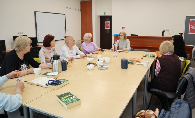 Na zdjęciu znajduje się osiem kobiet, które siedzą przy złączonych stołach. Przed kobietami leżą książki, a na środku na stole proporczyk oraz termosy, szklanki. Warsztaty odbywają się w sali komputerowej.