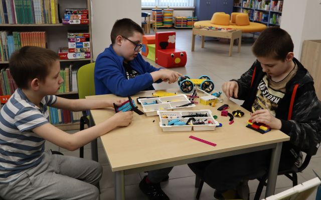 Na zdjęciu znajduje się trójka chłopców, którzy siedzą przy stoliku i układają wspólnie znajdujące się na stole klocki LEGO. W tle znajdują się półki z książkami.