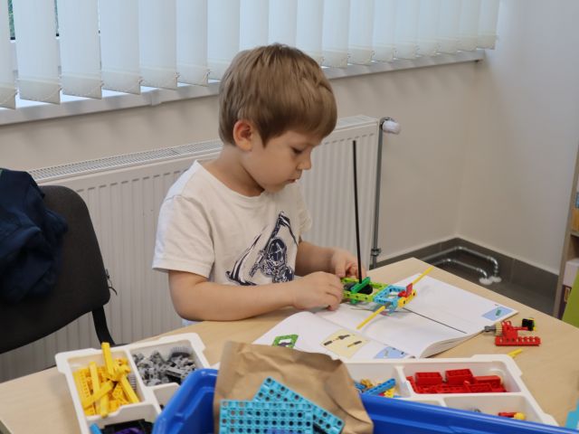 Na zdjęciu znajduje się chłopiec, który układa klocki LEGO przy stoliku. W tle znajdują się okna.