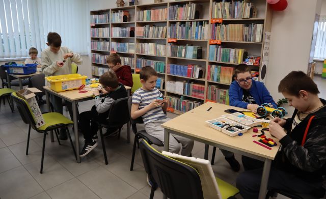 Na zdjęciu znajduje się siedmiu chłopców przy trzech stolikach, przy których układają klocki LEGO. W tle znajdują się półki z książkami.