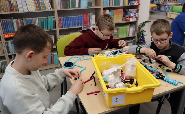 Na zdjęciu znajduje się trzech chłopców, którzy układają klocki LEGO przy stoliku, na którym leży plastikowe pudełko. W tle znajdują się półki z książkami.
