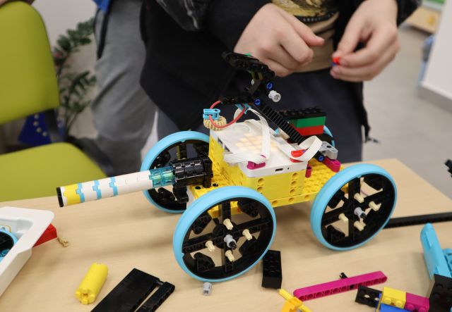 Na zdjęciu znajduje się leżący na stole wokół klocków, zbudowany z LEGO samochód, za nim przy stoliku stoi chłopiec, który nie został uchwycony w całości.