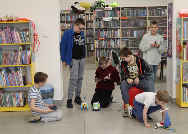 Na zdjęciu znajduje się sześciu chłopców, czterech z nich kuca, dwóch na podłodze prze nimi leżą trzy zbudowane z klocków LEGO pojazdy. W tle znajdują się półki z książkami.