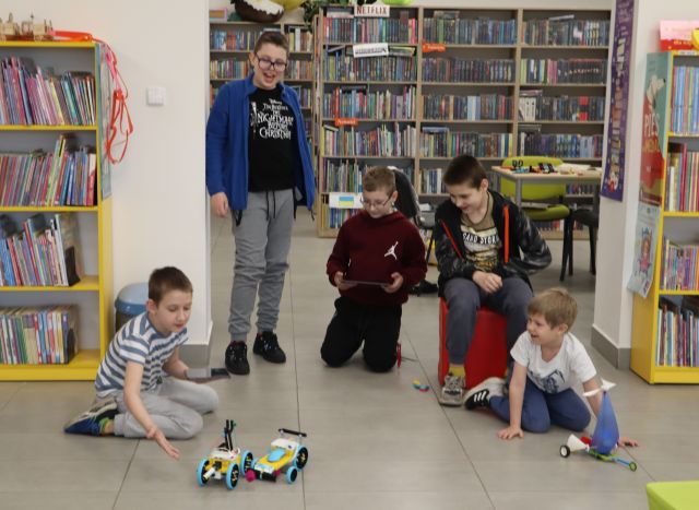 Na zdjęciu znajduje się pięciu chłopców, czterech z nich kuca. Przed nimi znajdują się trzy pojazdy z klocków, jeden po prawej stronie, a dwa po lewej obok siebie. W tle znajdują się półki z książkami.