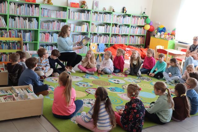 Na zdjęciu znajduje się siedząca na dywanie w okręgu grupa przedszkolaków, na środku między nimi siedzi kobieta z otwartą książką. W tle znajdują się półki z książkami.