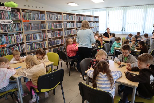 Na zdjęciu znajdują się przedszkolaki przy pięciu stolikach. Pomiędzy stolikami chodzi kobieta. W tle znajdują się półki z książkami.