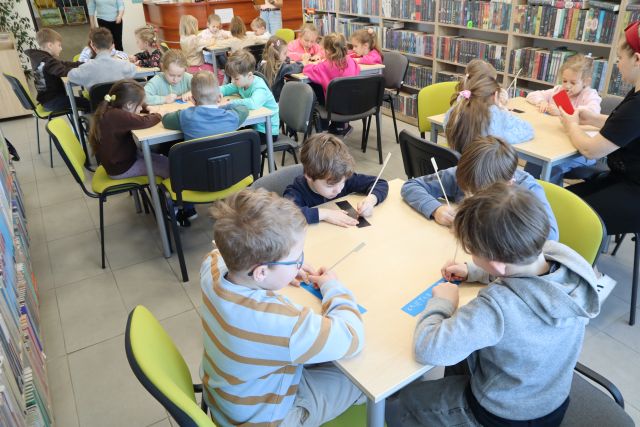 Na zdjęciu znajdują się przedszkolaki przy stolikach i robią swoje zakładki do książek. Po prawej stronie znajdują się półki z książkami.
