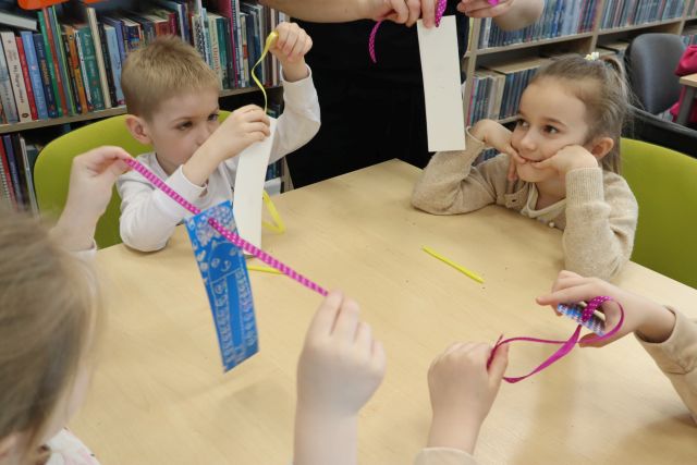 Na zdjęciu znajduje się czwórka przedszkolaków przy stoliku i zakładają wstążki na swoje zakładki do książek. W tle znajdują się półki z książkami.