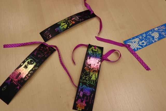 Na zdjęciu znajdują się leżące na stole cztery zakładki wykonane przez dzieci z wydrapywanki, posiadają wstążki.