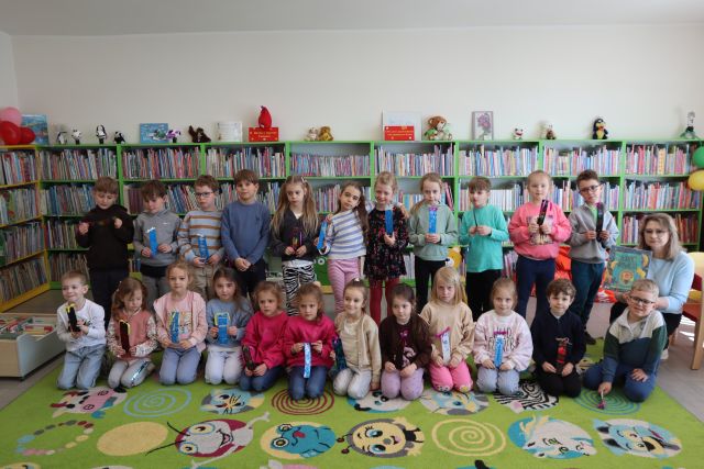 Na zdjęciu znajdują się przedszkolaki ustawione do zdjęcia, część z nich kuca na dywanie, po prawej stronie przy dzieciach kuca kobieta z książką. W tle znajdują się półki z książkami.