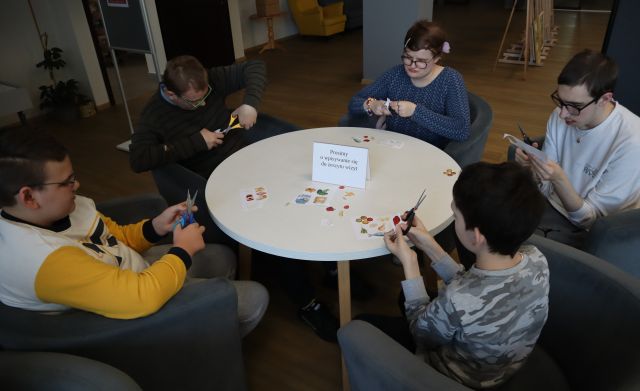 Na zdjęciu znajduje się piątka młodzieży siedząca przy okrągłym stoliku i wycinają kolorowe produkty spożywcze z kartki. Warsztaty odbywają się na holu biblioteki.