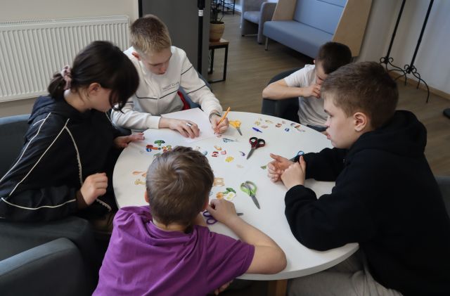 Na zdjęciu znajduje się piątka nastolatków przy okrągłym stole i wycinają kolorowe warzywa a kartek. Warsztaty odbywają się na holu biblioteki.