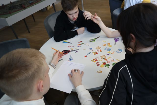 Na zdjęciu znajduje się trójka nastolatków przy stoliku, na którym leżą powycinane kolorowe obrazki, nożyczki oraz kartka. Warsztaty odbywają się na holu biblioteki.