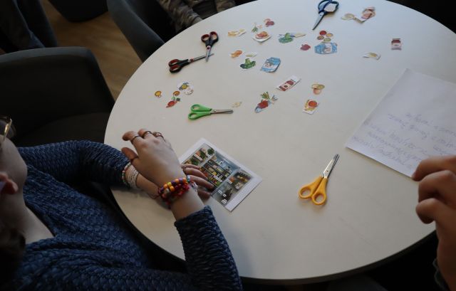 Na zdjęciu znajduje się stolik, na którym znajdują się powycinane owoce i warzywa, nożyczki i kartki. Z lewej strony przy stole siedzi kobieta ale nie została cała uchwycona na zdjęciu.