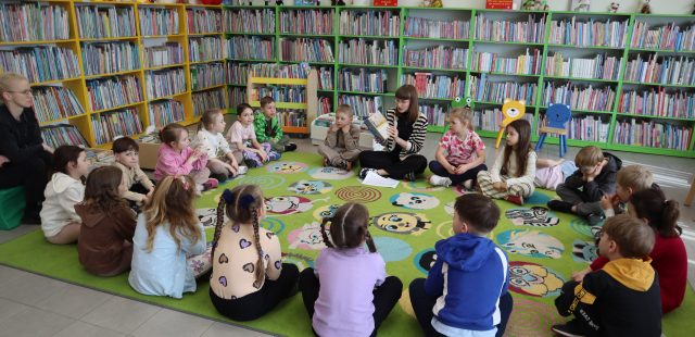 Na zdjęciu znajduje się grupa przedszkolaków siedząca w okręgu na dywanie, po środku siedzi kobieta z książką. W tle znajdują się półki z książkami.