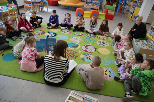 Na zdjęciu znajduje się grupa przedszkolaków siedząca na dywanie w okręgu wraz z kobietą, która trzyma w ręce książkę. W tle znajdują się pufy ułożone w pociąg.