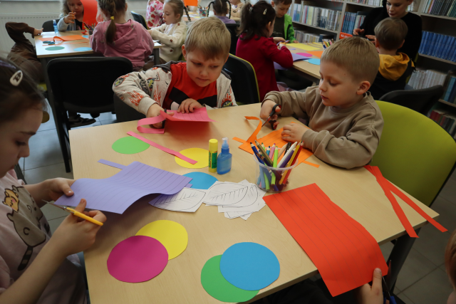 Na zdjęciu znajdują się przedszkolaki przy stoliku i wycinają paski z kolorowych kartek, na stoliku leżą również kolorowe kółka, kredki i kleje. W tle znajduje się reszta przedszkolaków przy stolikach.