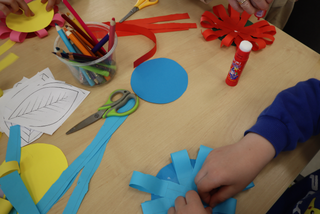 Na zdjęciu znajduje się stolik z kredkami, nożyczkami i kolorowymi kartkami, na stoliku leżą również dwa wykonane z papieru kwiaty.