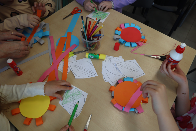 Na zdjęciu znajduje się stolik z kolorowymi kartkami, wydrukowanymi liśćmi oraz dziewczynki, których ręce zostały uchwycone na zdjęciu, kleją i kolorują swoje prace.