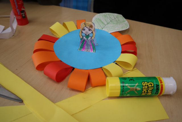 Na zdjęciu znajduje się wykonana z papieru Calineczka na kolorowym papierowym kwiecie, przed pracą leżą żółte kartki i klej.