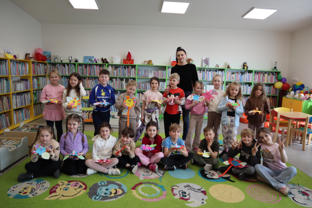 Na zdjęciu znajduje się dziewiętnaście przedszkolaków, część siedzi, a część stoi do zdjęcia ze swoimi pracami, za nimi stoi kobieta. W tle znajdują się półki z książkami.