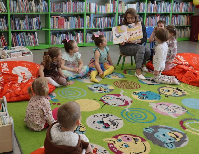 Na zdjęciu znajdują się dzieci na dywanie wraz z kobietą, która pokazuje im ilustrację w książce. Po lewej i prawej stronie leżą czerwone pufy, a w tle znajdują się półki z książkami.
