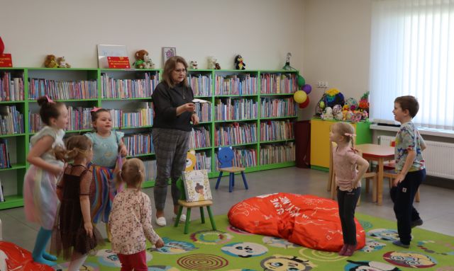 Na zdjęciu znajdują się stojące na dywanie dzieci wraz z kobietą, na dywanie leży czerwona pufa. W tle znajdują się półki z książkami.