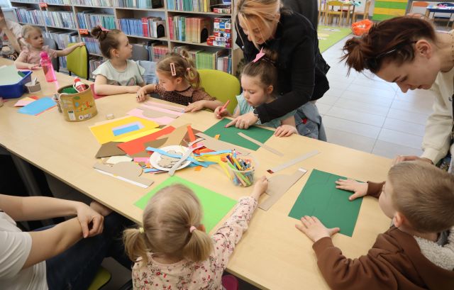 Na zdjęciu znajdują się dzieci wraz z rodzicami, które przy stolikach wycinają z kolorowych kartek ozdoby wróżkowe. W tle znajdują się półki z książkami.