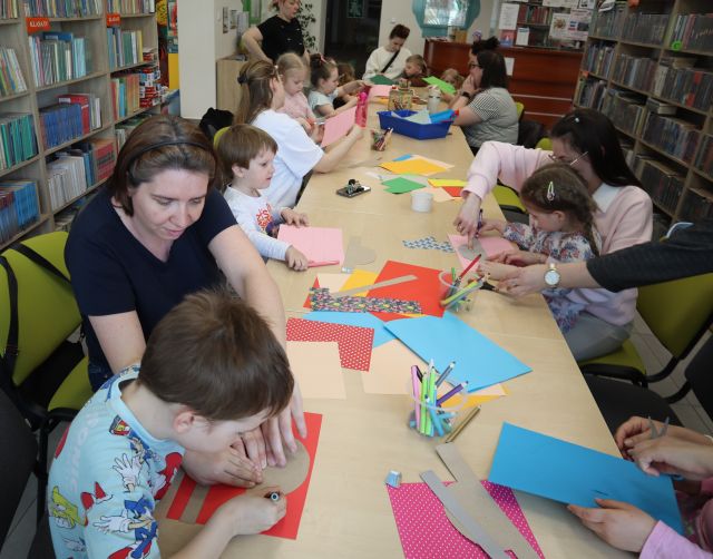 Na zdjęciu znajduje się stół z kolorowymi kartkami, kredkami, a dookoła siedzą rodzice z dziećmi/ W tle i po bokach znajdują się półki z książkami.