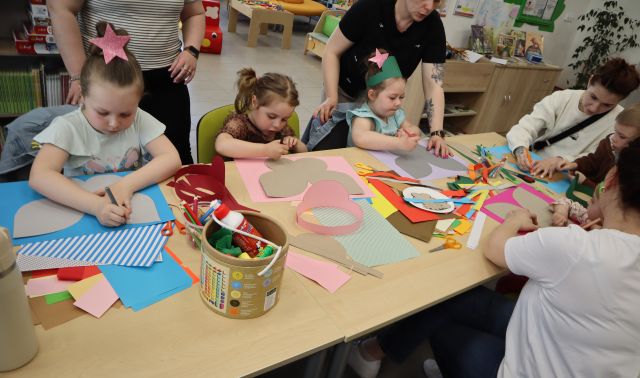 Na zdjęciu znajdują się dzieci odrysowujące skrzydła na kolorowych kartkach, które leżą na stole, obok dzieci znajdują się rodzice. W tle znajdują się szafki.