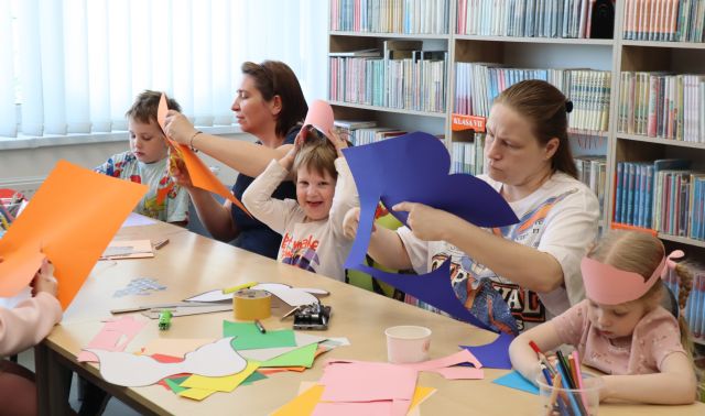 Na zdjęciu znajdują się dzieci z rodzicami i wycinają i skrzydła i korony z kolorowych kartek, leżących na połączonych stołach. W tle znajdują się półki z książkami.