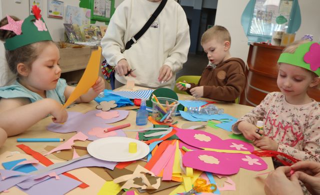 Na zdjęciu znajduje się trójka dzieci, dwie dziewczynki mają na głowie papierowe korony, a na stole przed nim leżą kolorowe kartki i skrzydła. Przy chłopcu po środku stoi kobieta. W tle znajdują się szafki, obrazki na ścianach.