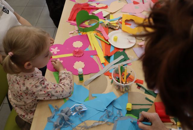 Na zdjęciu znajdują się połączone stoły, na których leżą kolorowe kartki oraz skrzydła, do których dziewczynka dokleja kwiatki.