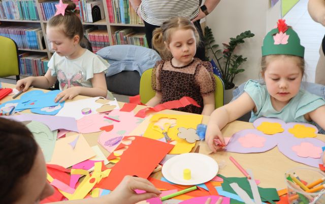 Na zdjęciu znajdują się trzy dziewczynki , jedna ma na głowie papierową koronę. Dziewczynki ozdabiają swoje kolorowe skrzydła przy stoliku. W tle znajdują się półki z książkami.