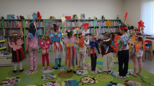 Na zdjęciu znajduje się dziesiątka dzieci stojąca tyłem do zdjęcia, na plecach mają skrzydła i podnoszą do góry trzymane w ręce różdżki. W tle znajdują się półki z książkami.