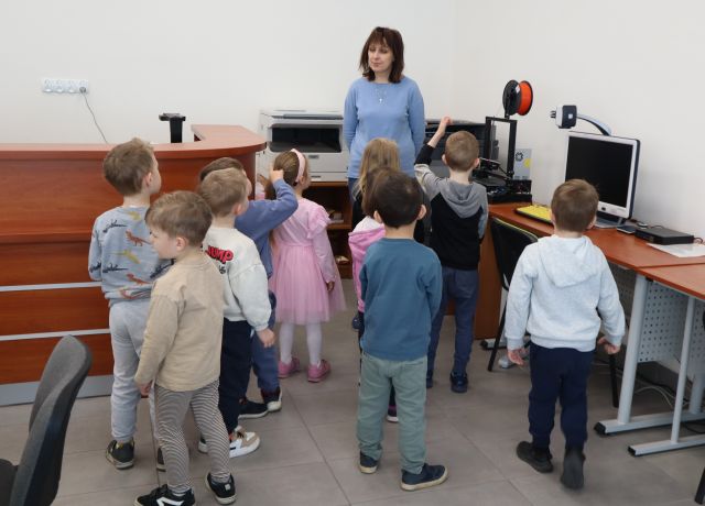 Na zdjęciu znajduje się grupa dziesięciu przedszkolaków wraz z kobietą w sali komputerowej, przy drukarce 3D.