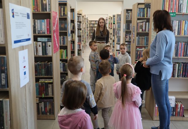 Na zdjęciu znajduje się grupa przedszkolaków idąca między półkami z książkami w bibliotece, przed nimi stoi jedna kobieta oraz po prawej stronie przy półkach także stoi kobieta.