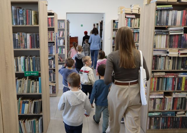 Na zdjęciu znajduje się dziesiątka dzieci idąca między półkami z książkami wraz z trzema kobietami. Zdjęcie zostało wykonane od tyłu.