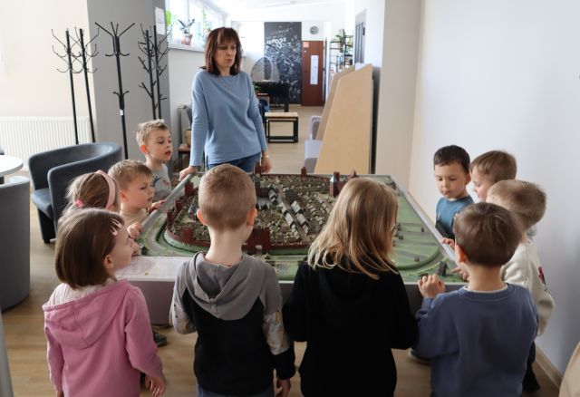 Na zdjęciu znajduje się kobieta oraz grupa przedszkolaków stojąca dookoła makiety miasta ze średniowiecza. W tle znajduje się kącik młodzieżowy, wieszaki oraz fotele po lewej stronie.